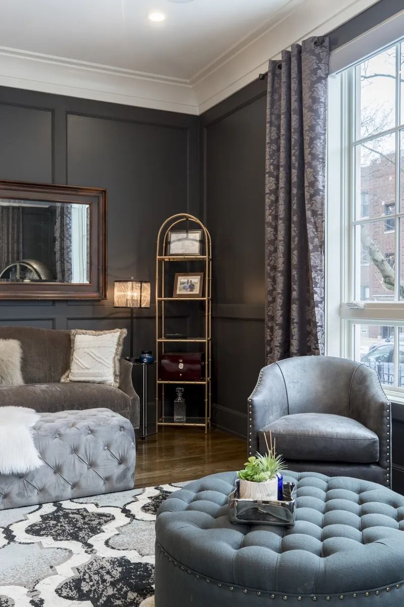 Elegant bedroom with dark paneled walls, tufted ottoman, gray chair, and large window with patterned curtains.