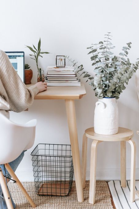 Desk with laptop, stacked books, plant, and white vase on a small stool in a bright minimalist room
