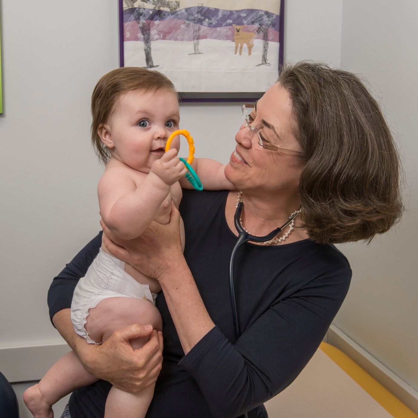 Doctor holding a baby with a teething ring, smiling. Doctor has a stethoscope on.