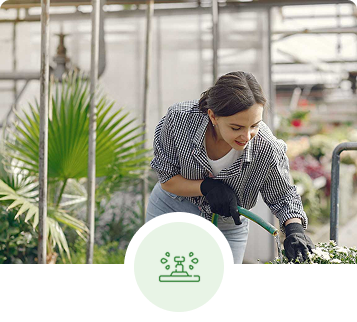 Woman watering plants in a greenhouse, smiling, wearing gloves.