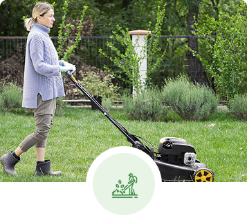 Woman mowing a lawn in a backyard. She wears a blue sweater, gloves, and boots.