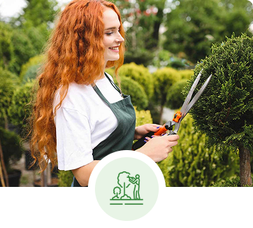 Woman with red hair, in green apron, smiles while trimming a bush with large shears in a garden.