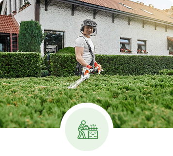 Man trimming hedges with a hedge trimmer, wearing protective gear, in front of a house.