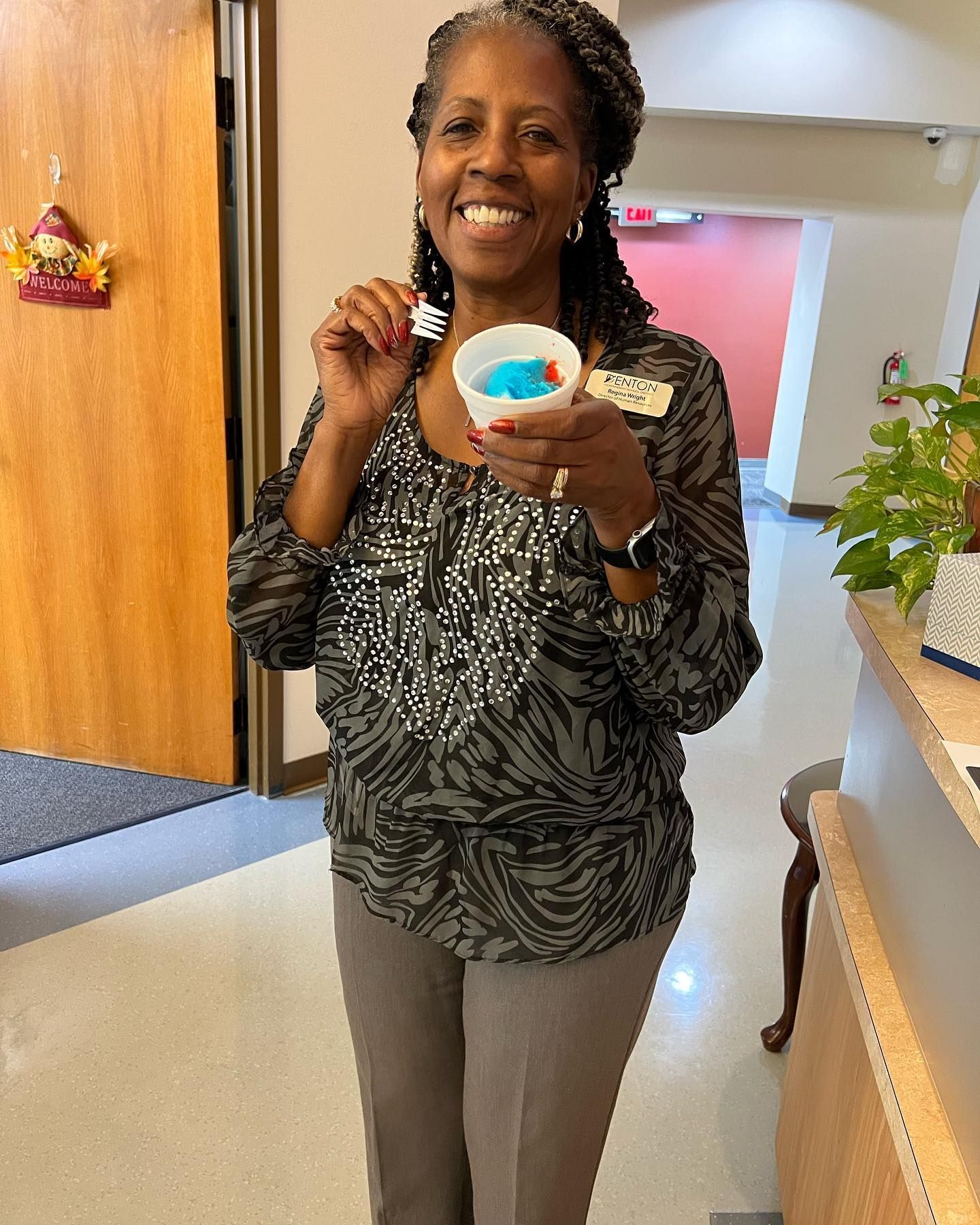 Woman smiling, holding a snow cone. Wearing a patterned top and gray pants. Indoors with a reception desk.