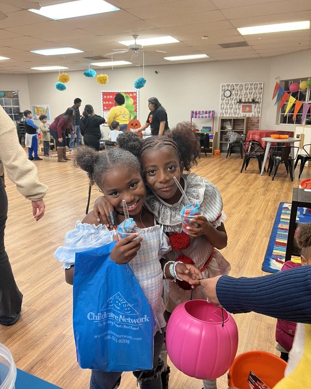 Two girls in costumes at an event, holding treats. One has a blue bag, other a pink pail.