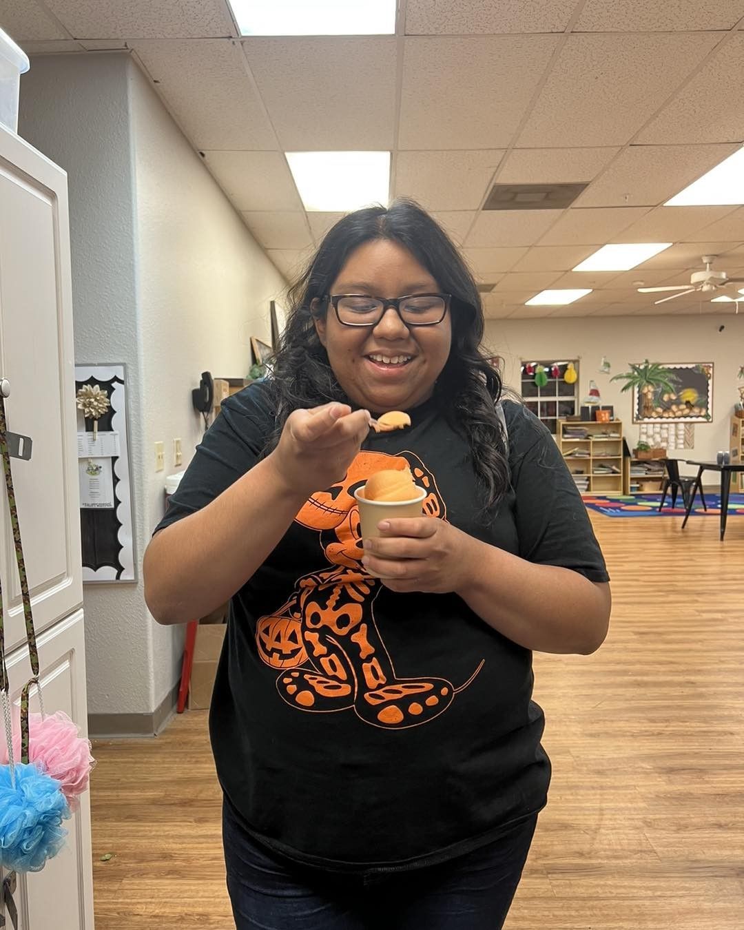 Woman smiling, holding a spoon over a small container. Wearing glasses and a black t-shirt with orange design. Indoors.