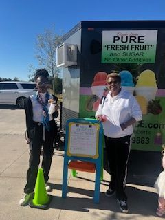 Two people stand beside a food truck. The truck has signs for fresh fruit. One person is holding a drink.