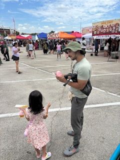 Man holding cotton candy, handing one to a child in a market. Blue sky, colorful tents, and people in the background.
