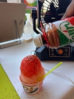 A person topping a snow cone with red chili powder from a bottle; indoors near a fan.