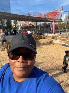 Woman in sunglasses and cap smiles, blue shirt. Outdoors: playground, people, red net structure.
