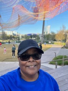 Woman wearing sunglasses and a cap smiles outside near public art and buildings.