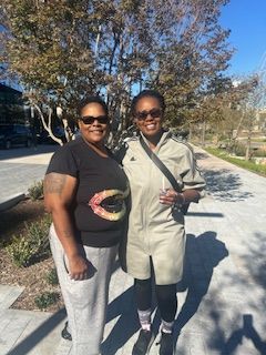 Two people standing side-by-side, outside on a sunny day. One wears a jumpsuit, the other a graphic tee.