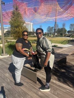 Two people outdoors, one with arm tattoo, holding a small object. Wooden deck, wire art above. Sunny day.