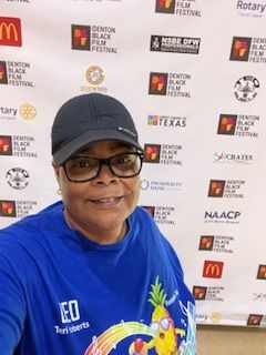Woman in blue shirt and cap smiles in front of a Denton Black Film Festival backdrop.