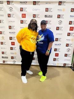 Two people pose in front of a banner with logos. One in yellow hoodie, one in blue shirt, both in black pants.