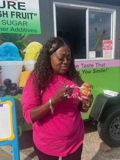 Woman in pink shirt eating snow cone in front of a food truck with colorful shave ice flavors.