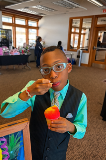 Boy in vest and tie eating a snack, indoors, holding a spoon, looking at the camera.
