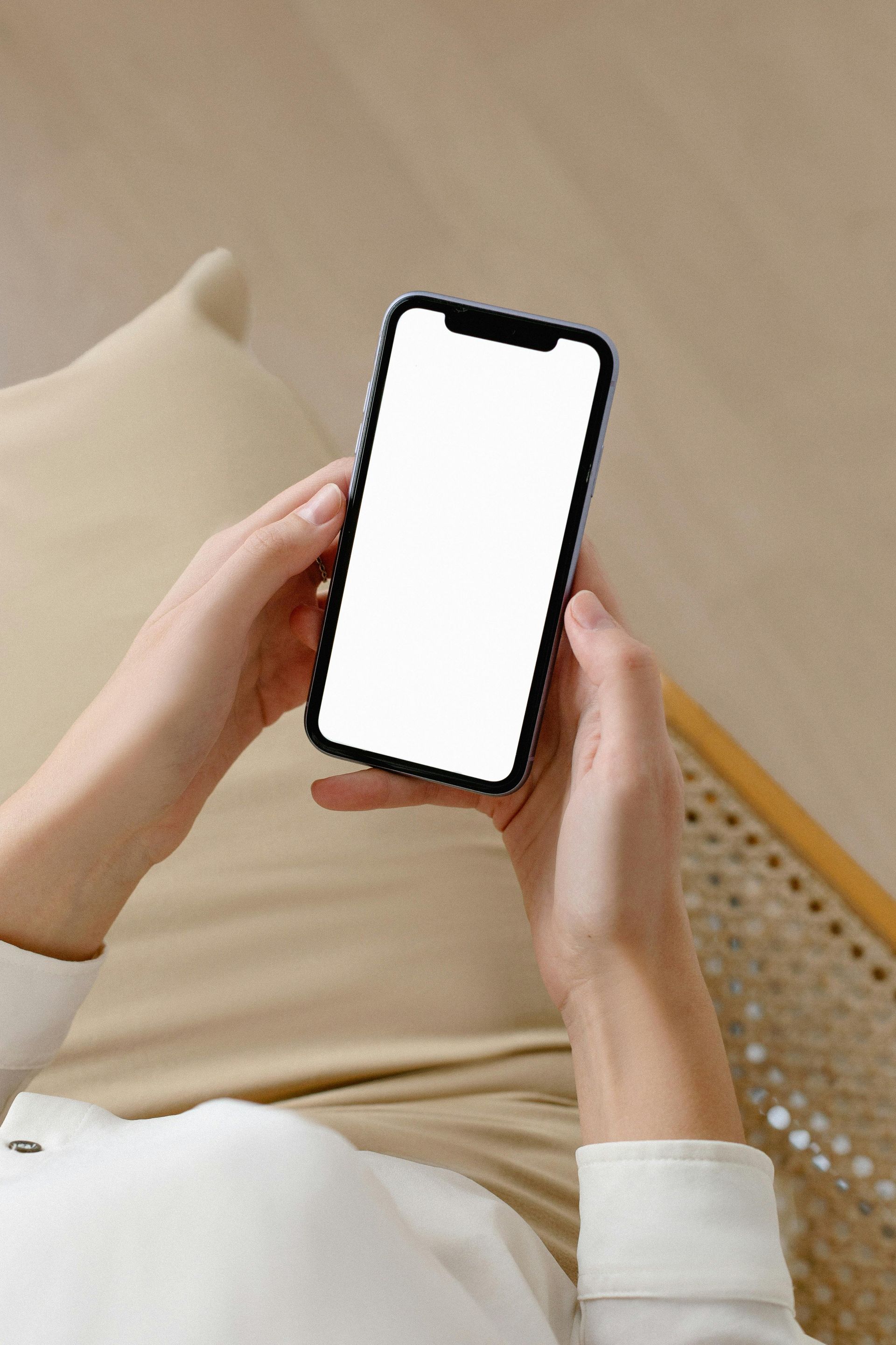 A woman is sitting on a couch holding a cell phone with a white screen.