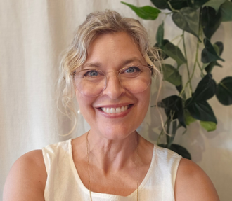 Smiling person in a white top with glasses, standing indoors beside a leafy plant.