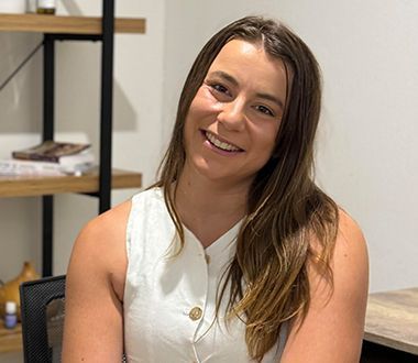 A woman in a white tank top is smiling for the camera while sitting at a desk.