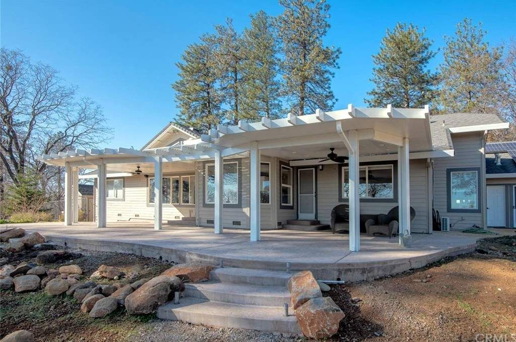 House with patio and pergola, steps down to a rocky area, trees in background, sunny day.