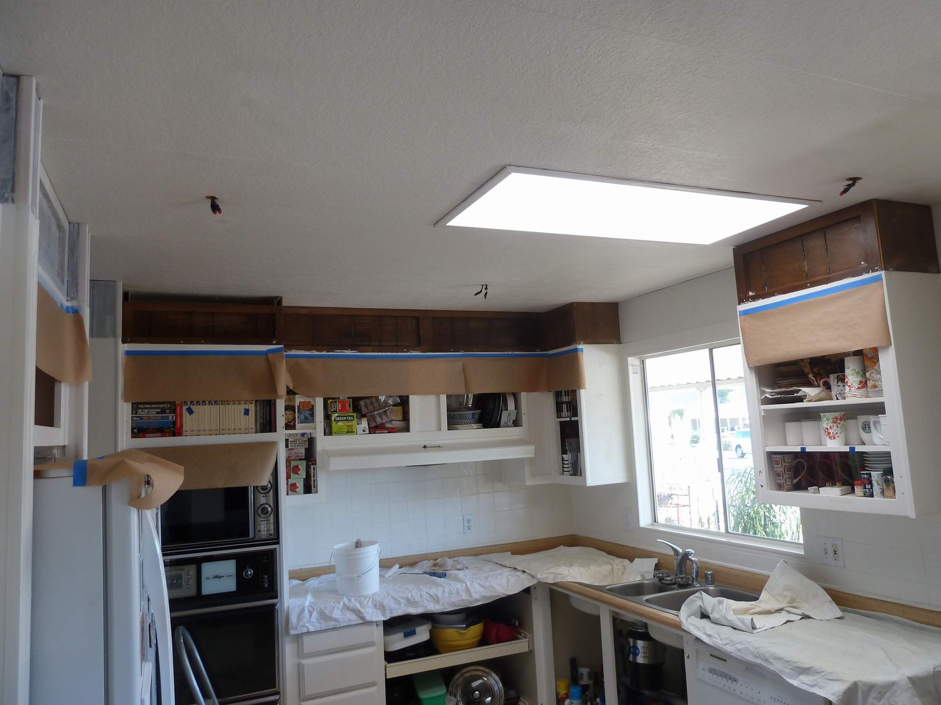 Kitchen interior being painted; cabinets and ceiling are masked. Brown upper cabinets, white lower cabinets.