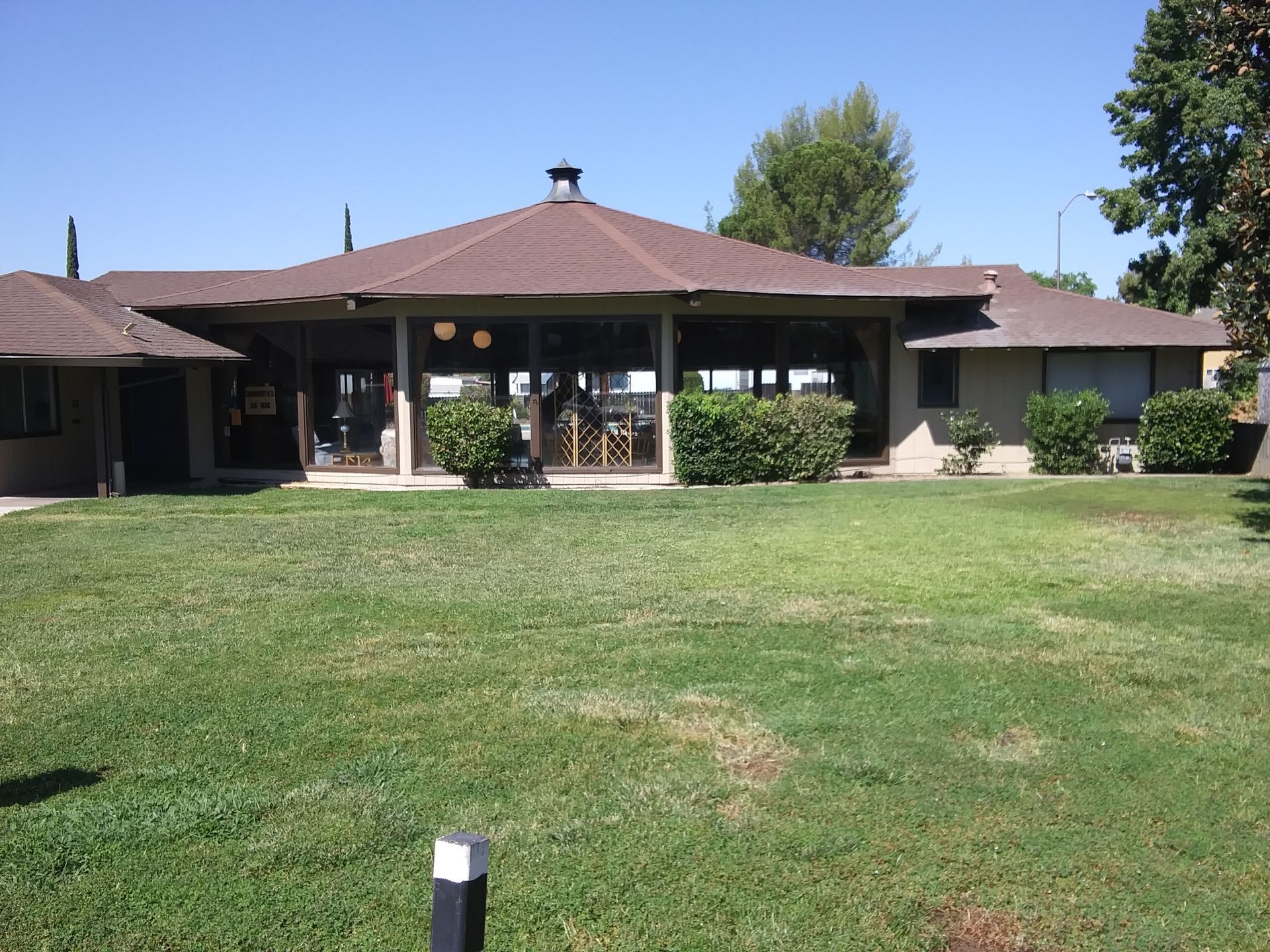 Brown-roofed building with glass walls, surrounded by green lawn and bushes, on a sunny day.