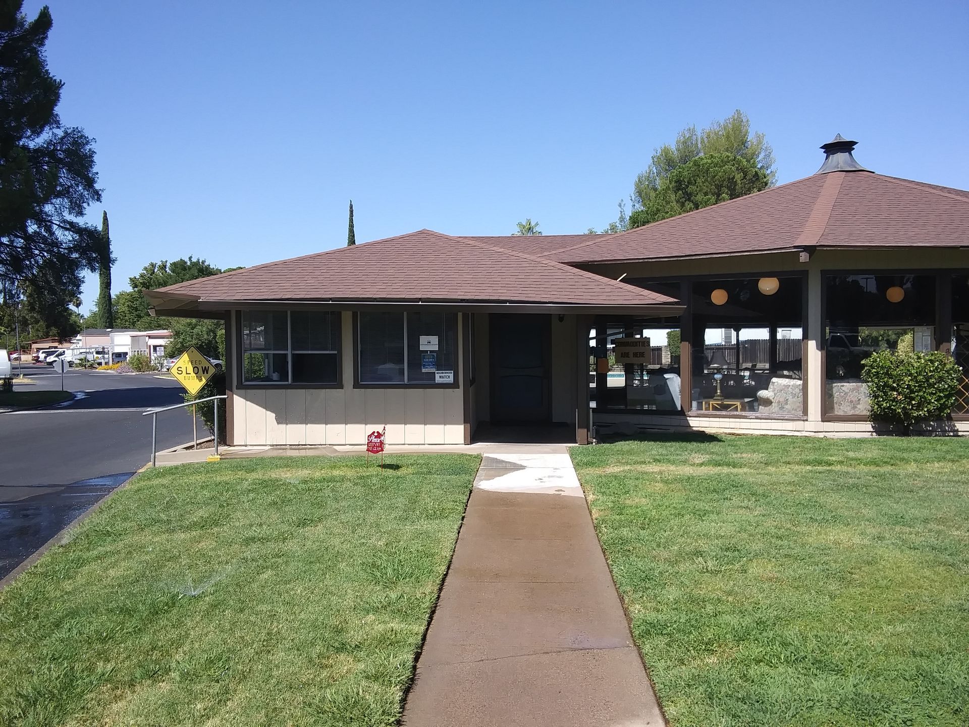 Lawn and walkway lead to a small beige building with a dark brown roof and an open-air gazebo.