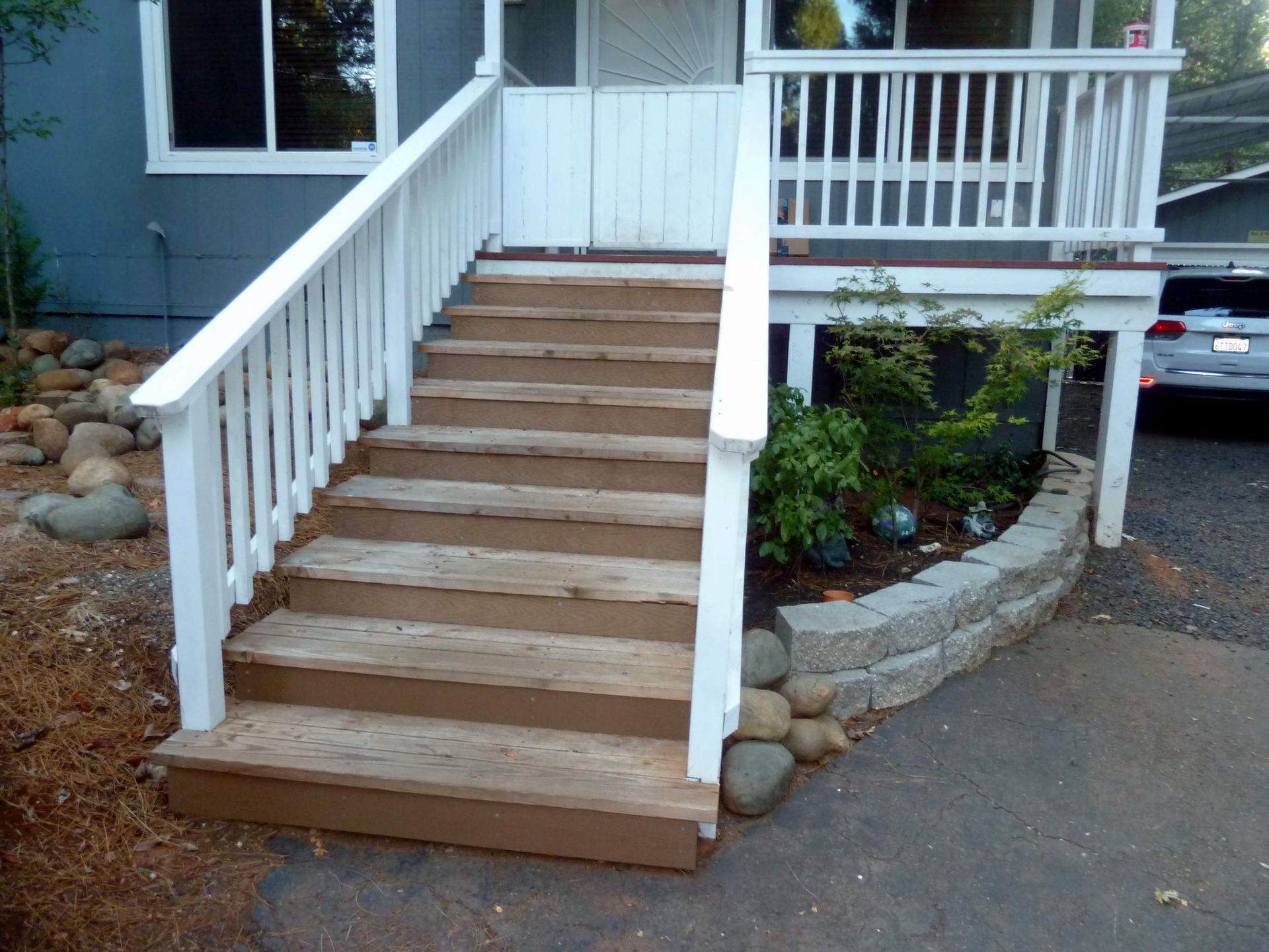 Wooden stairs with white railing leading to a gray house porch; stone retaining wall with greenery on the side.