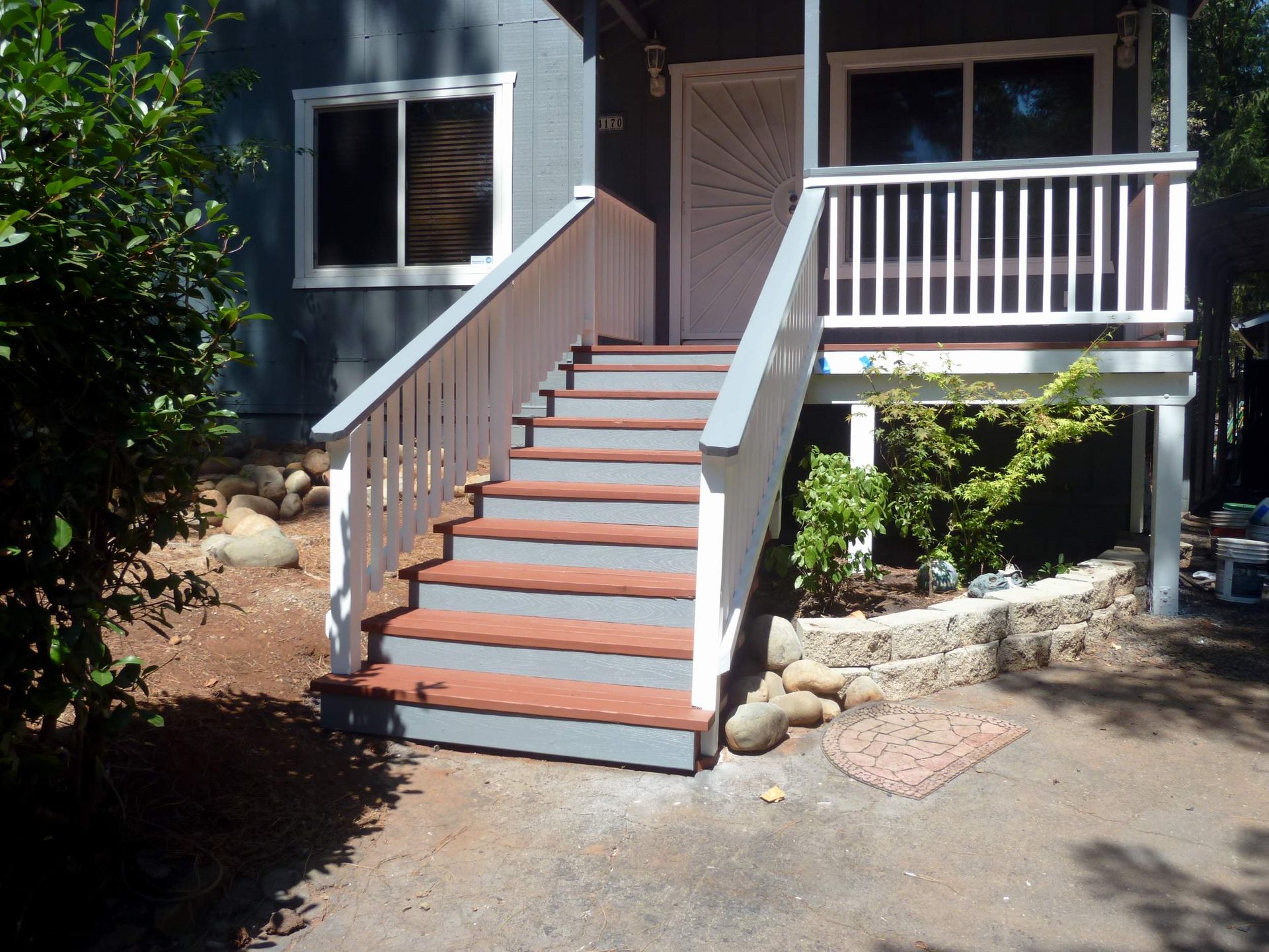 Gray house entrance with steps and white railing. Stone retaining wall with plants.