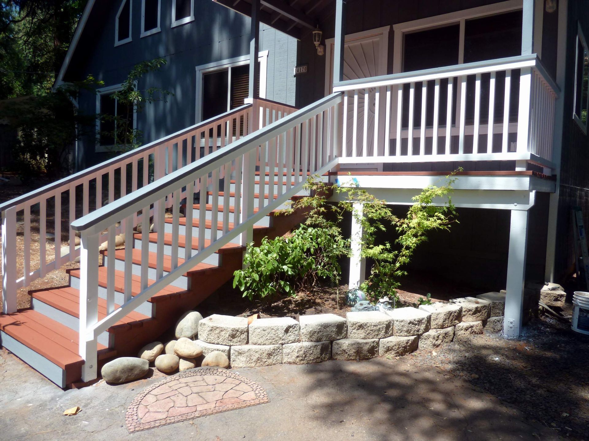 White-railed wooden staircase leading to a raised porch and front door of a gray house. Landscaping with stone wall at base.