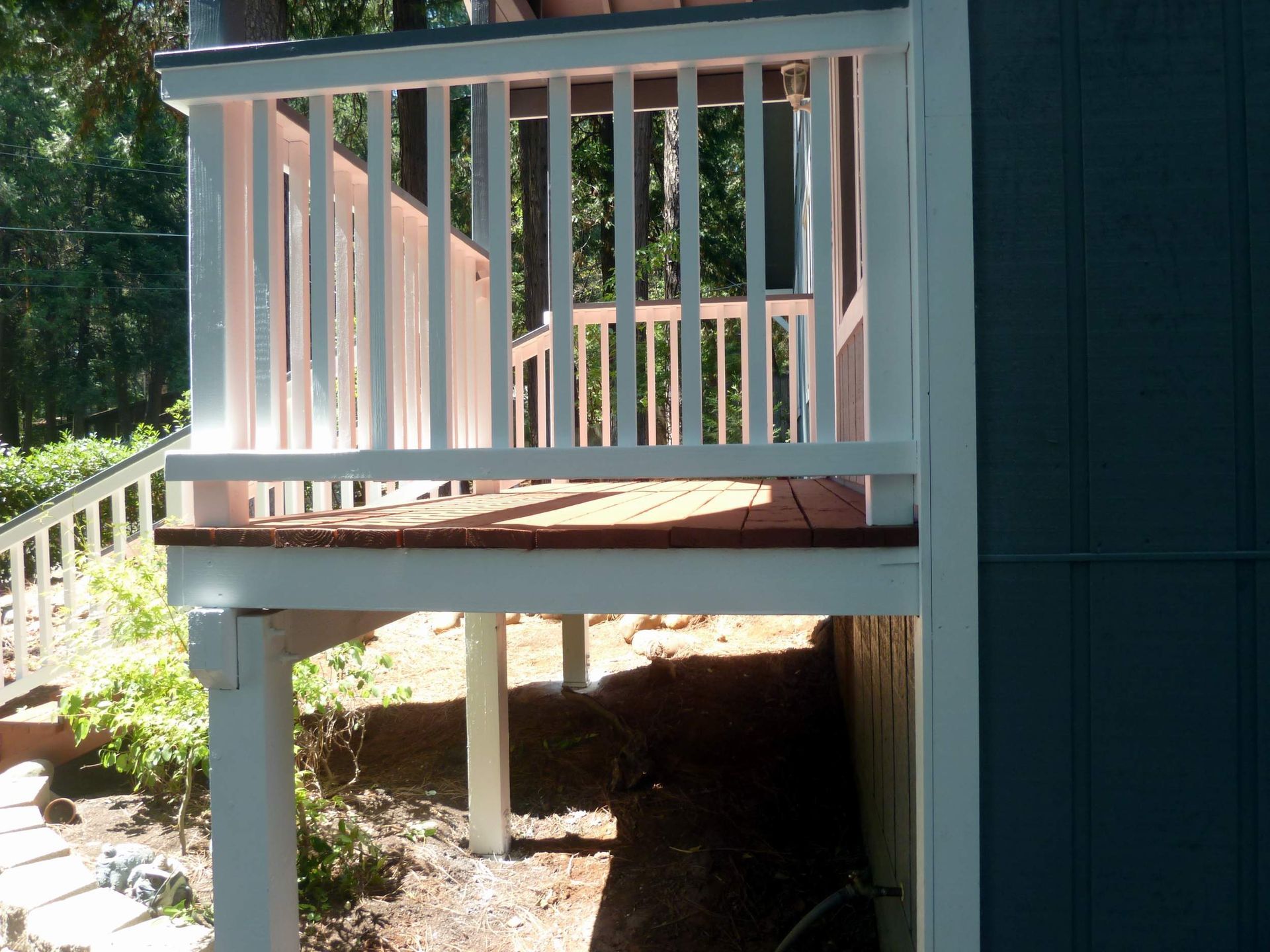White deck with railing attached to blue house, supported by white posts, with trees in the background.