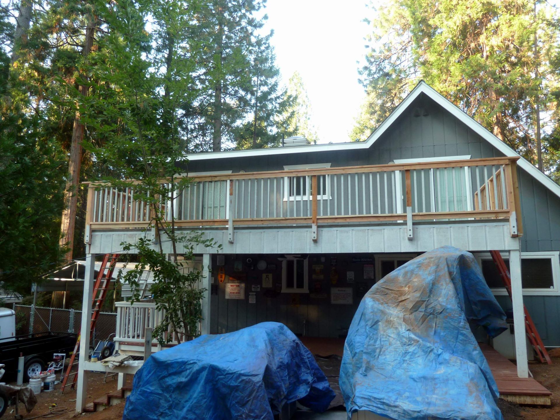 Two-story gray house with wooden deck, blue tarps covering materials, surrounded by trees.