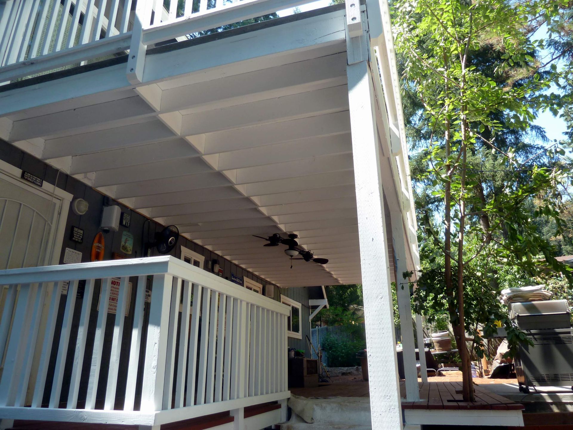 White painted deck with railing, beneath a second-story deck, trees on right.
