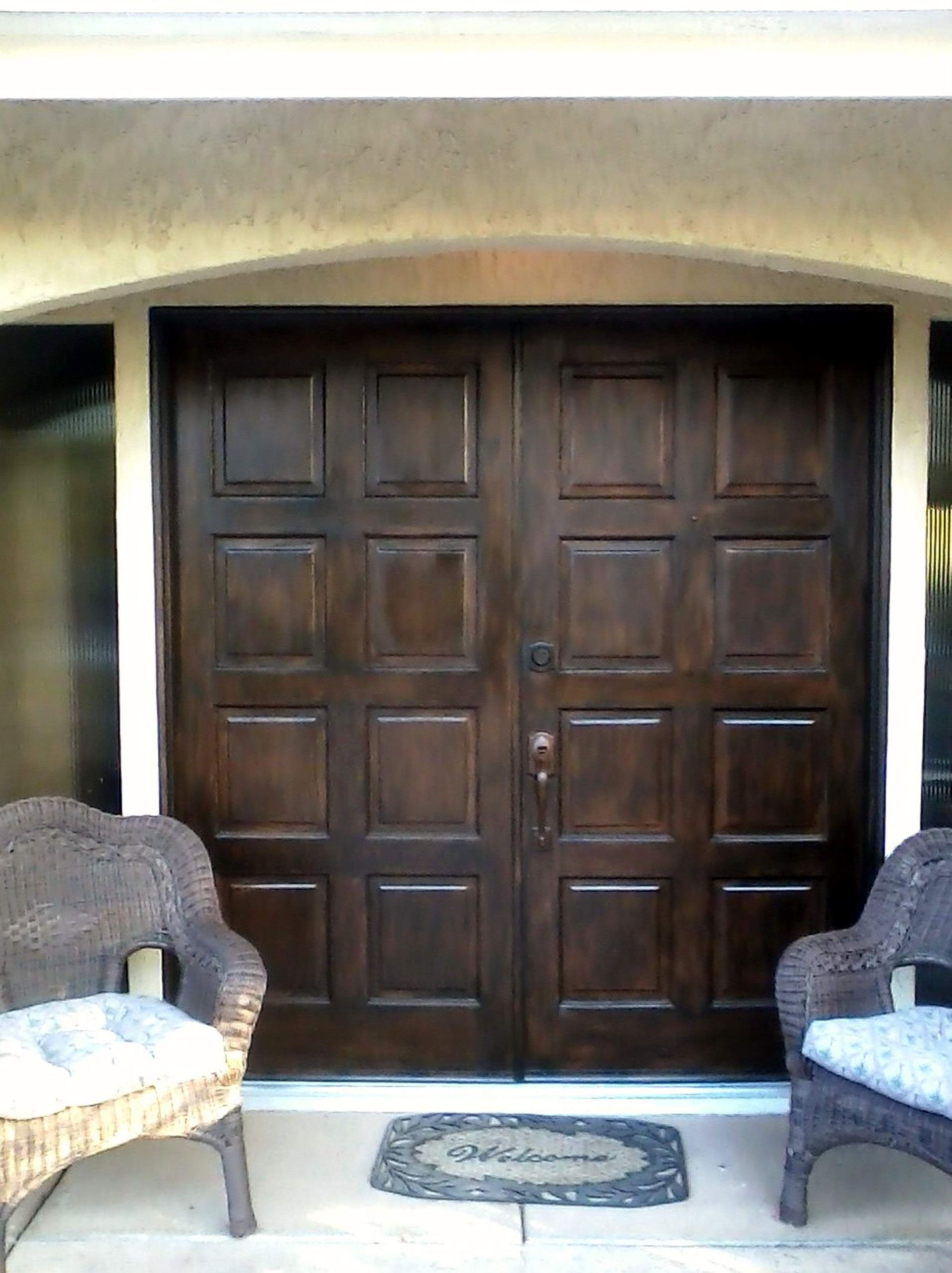 Brown double doors with paneled design, flanked by wicker chairs. A welcome mat rests on the porch.