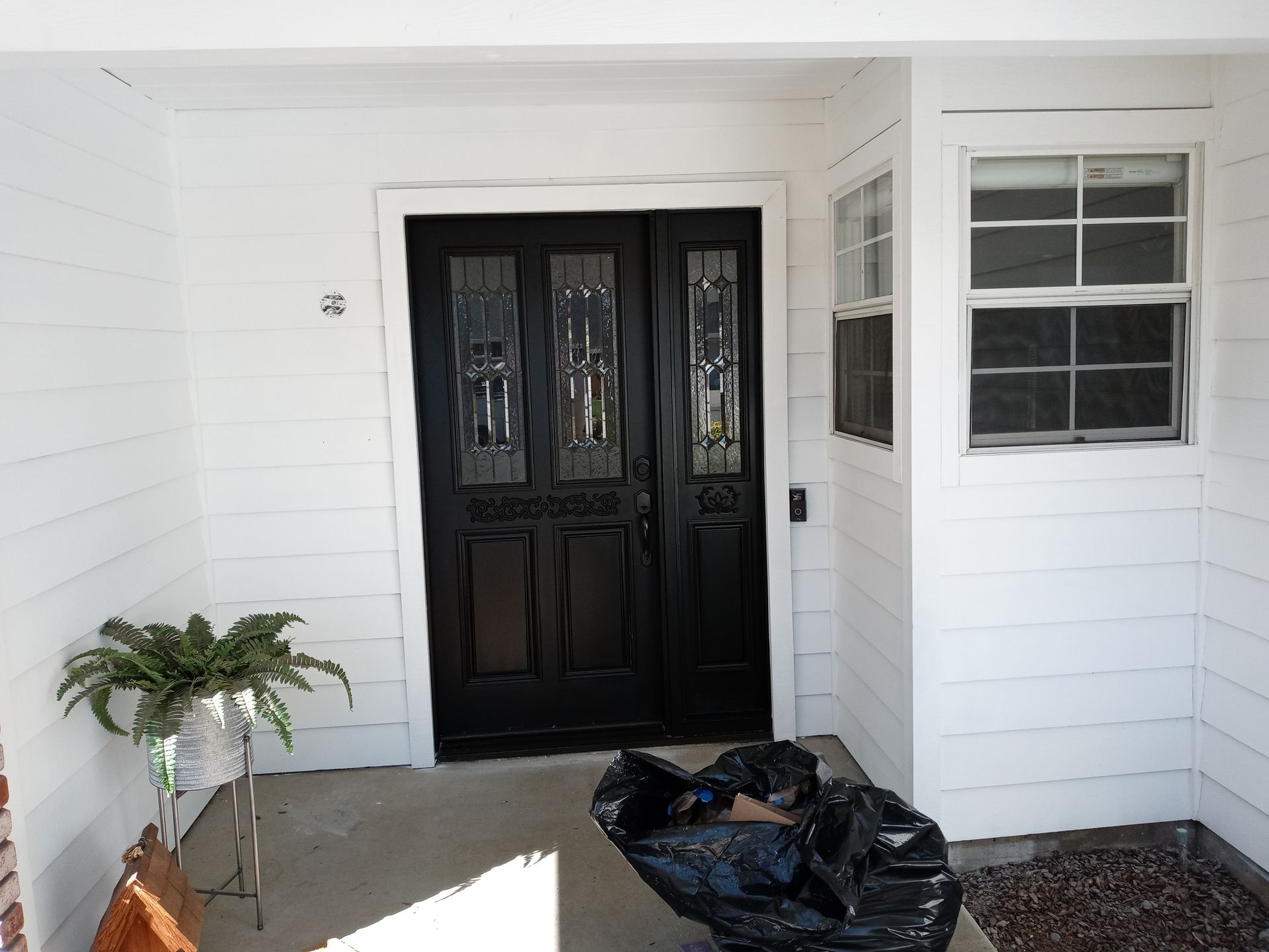 Black front door with glass panels on a white porch, potted plant to the left, trash bag in front.