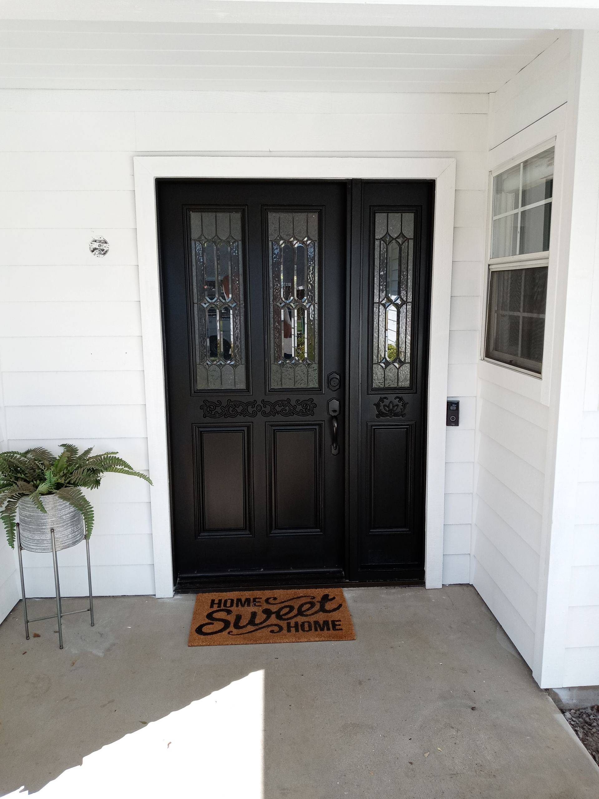 Black front door with glass panels and sidelight, beneath a white porch roof, a welcome mat, and a potted plant.
