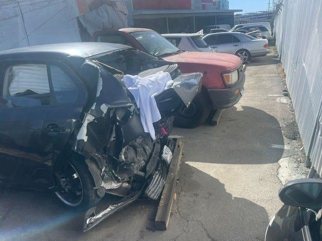 A severely damaged black car with a crumpled rear. A red truck is parked nearby.