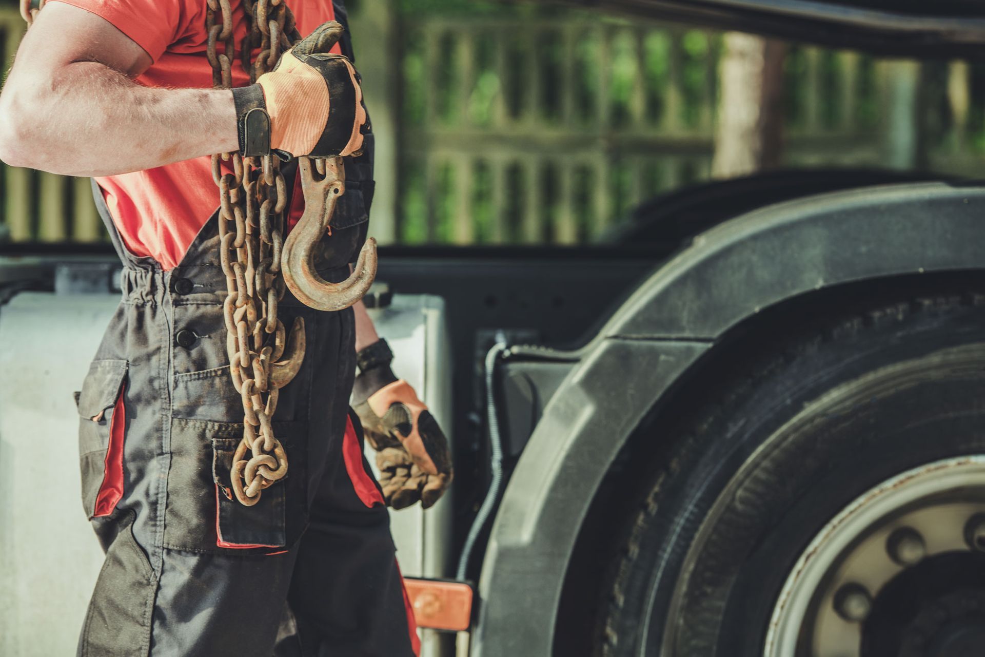 Tow truck operator holding chains near a truck's tire; wearing orange shirt, black overalls, and work gloves.