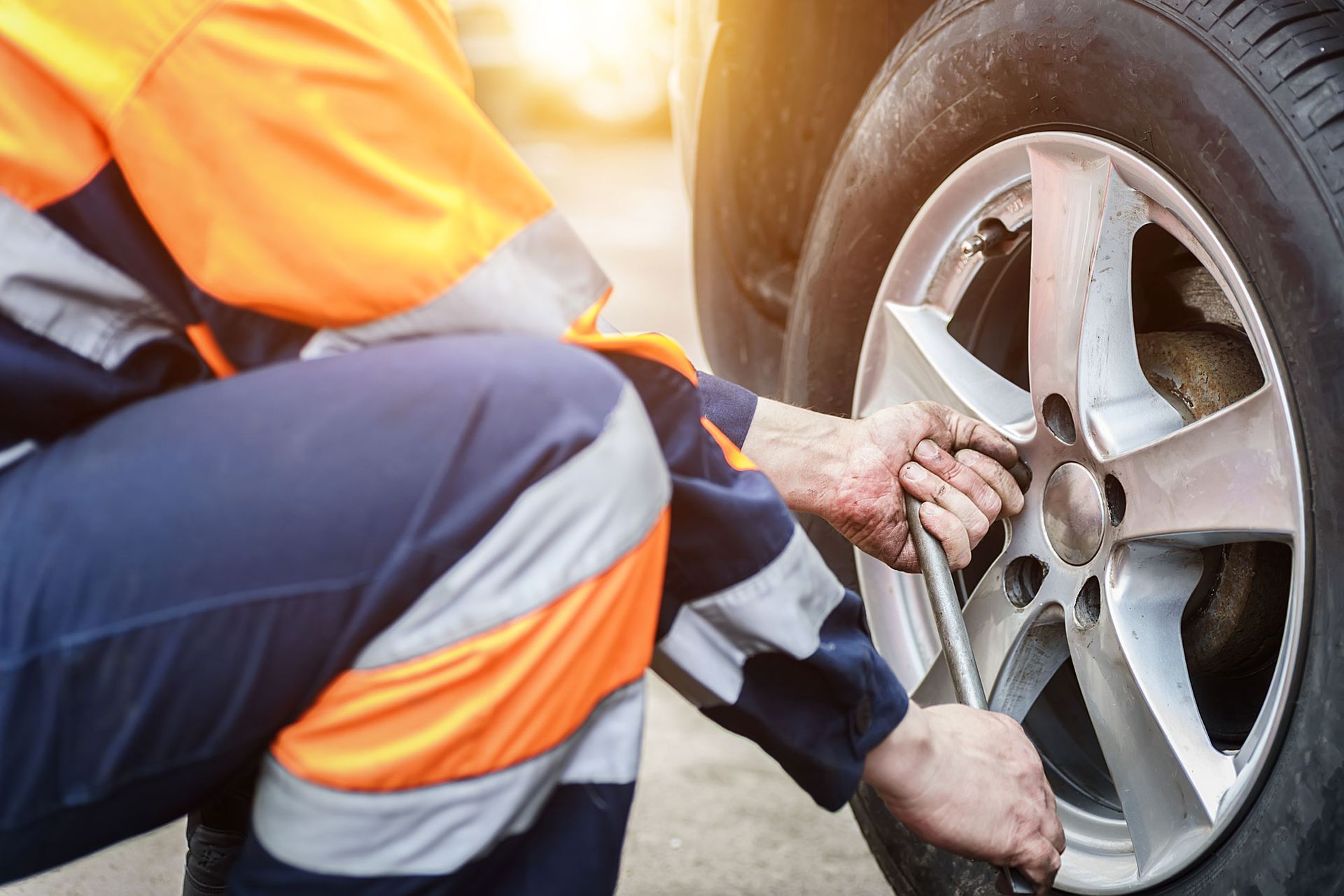 Mechanic in blue and orange overalls using a wrench to remove a car tire in an outdoor setting.