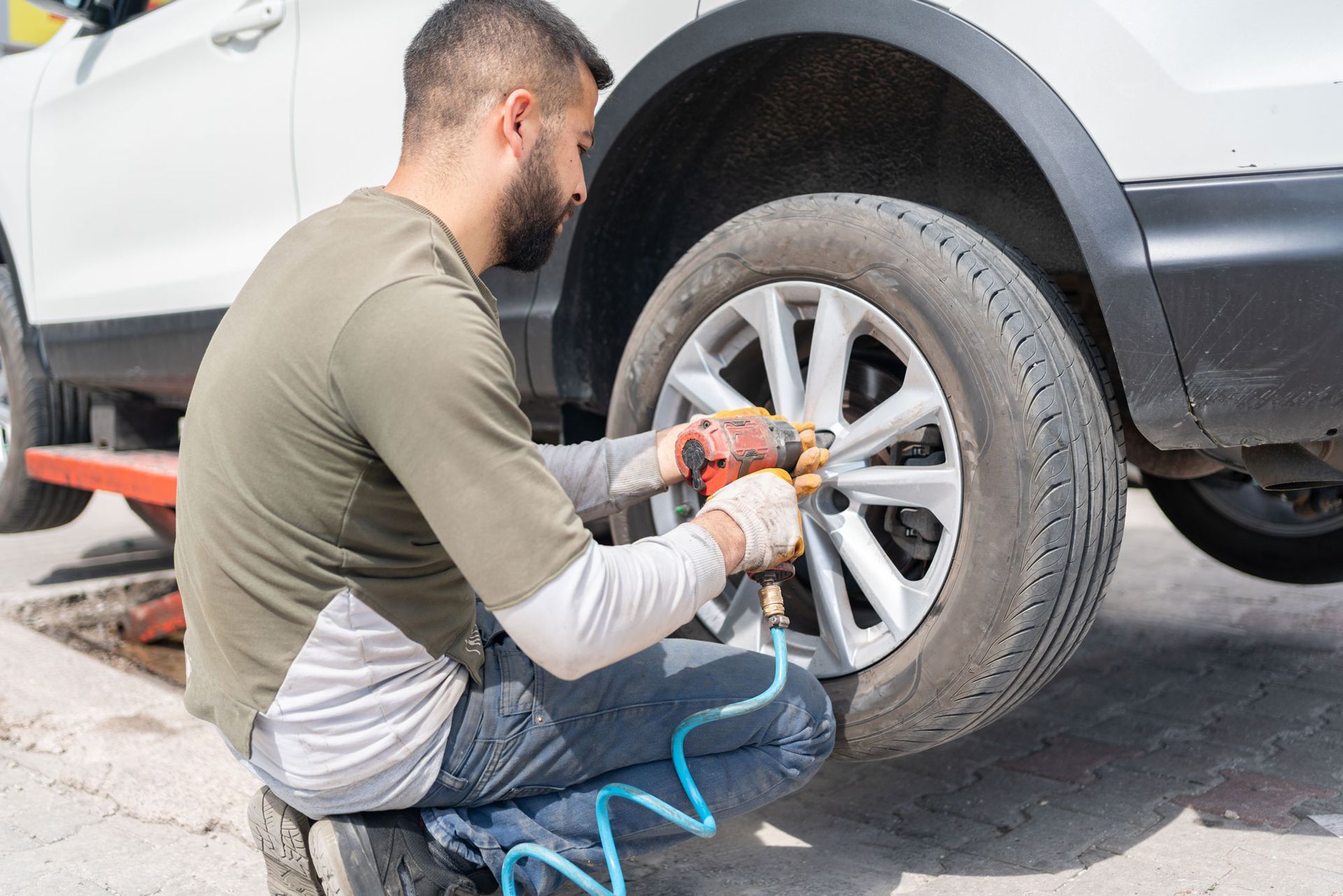 Man kneeling, using an impact wrench to remove lug nuts from a car tire, outdoors.