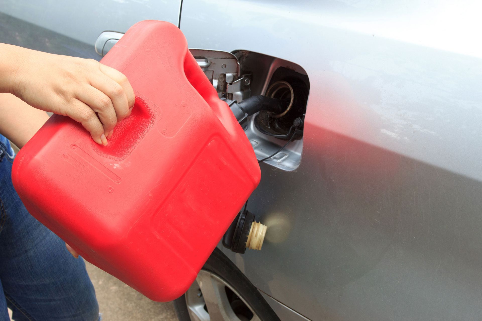 Person filling a car's gas tank with red gasoline container. Silver car, fuel door open.