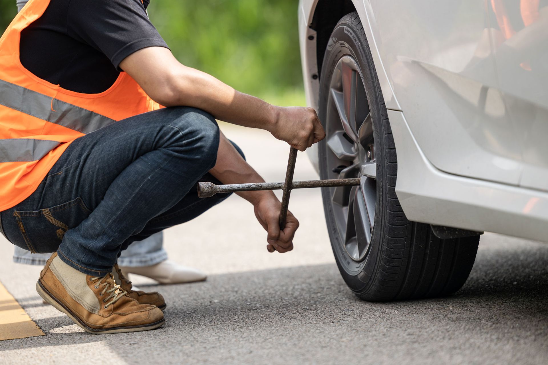 Person in an orange vest changing a flat tire on a white car with a lug wrench on the side of the road.