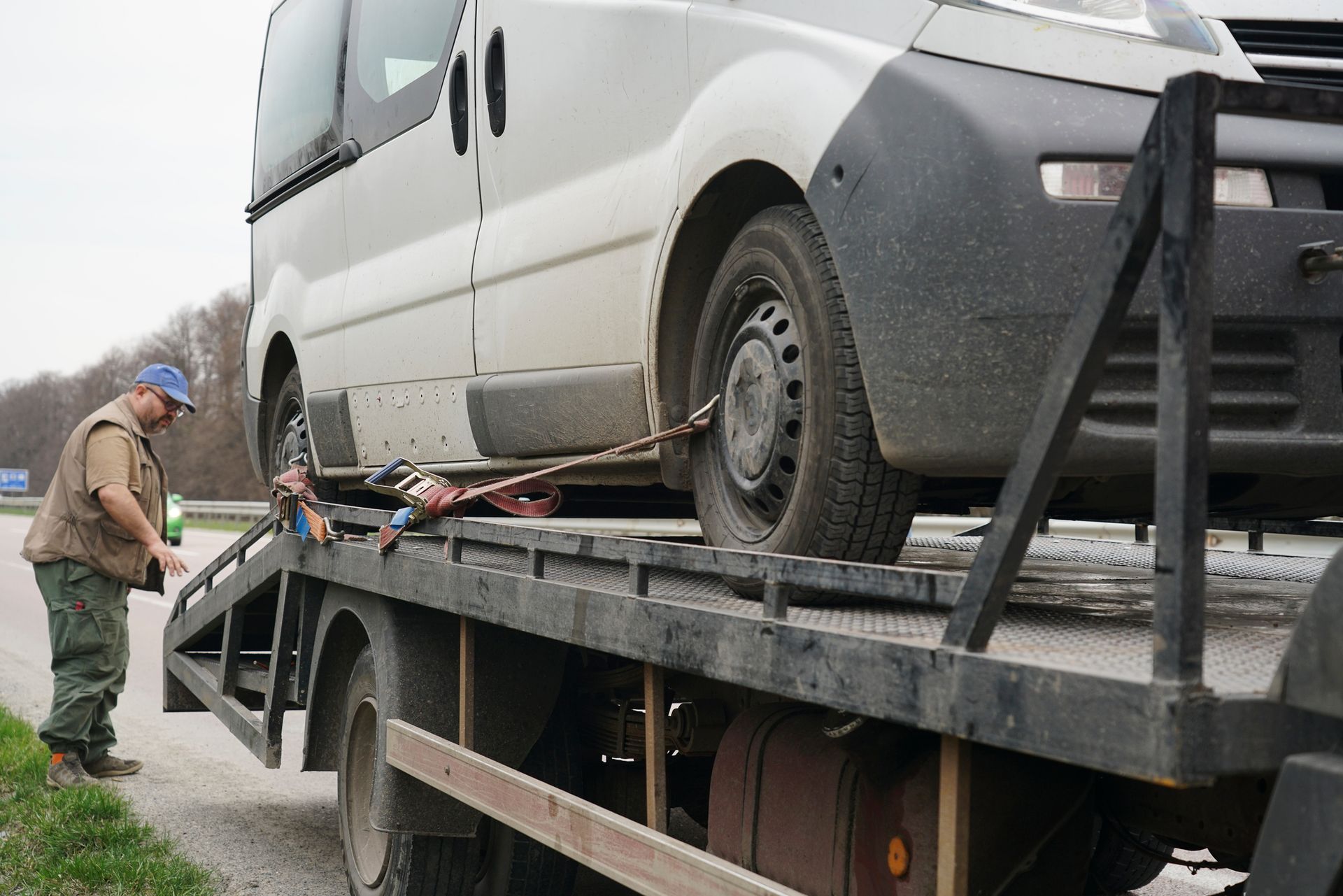 A van secured on a flatbed tow truck with straps along a roadside.