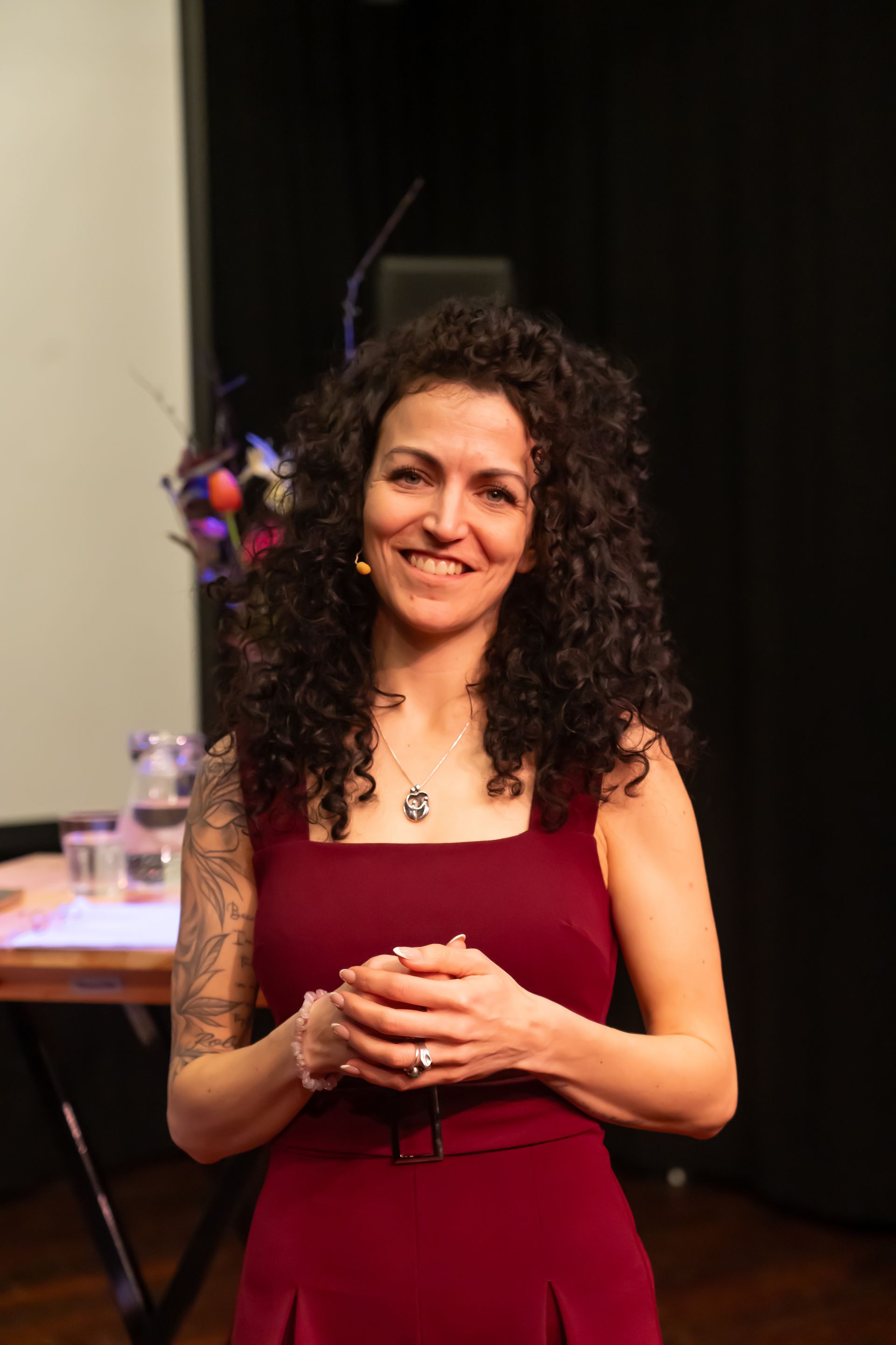 A person with curly dark hair and a tattoo on their arm, wearing a maroon sleeveless top, smiles in an indoor setting.