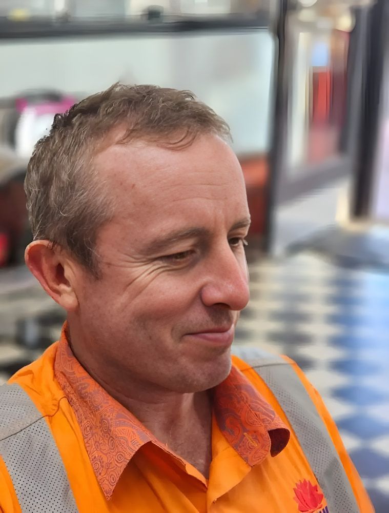 A Man Wearing An Orange Shirt And Safety Vest Is Smiling — Get It Off Men's Barber Shop In Raymond Terrace, NSW