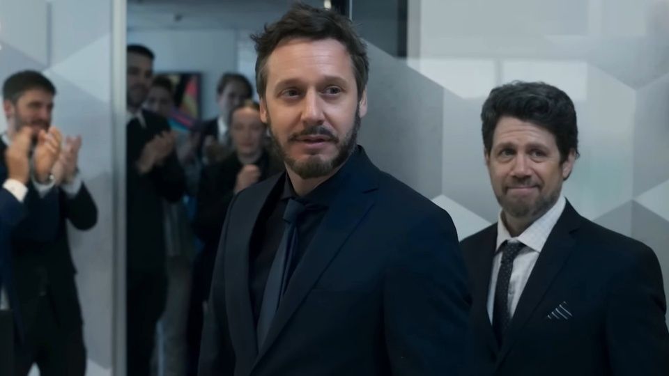 Un hombre de traje negro, seguido de otro, entra en una habitación mientras la gente aplaude.