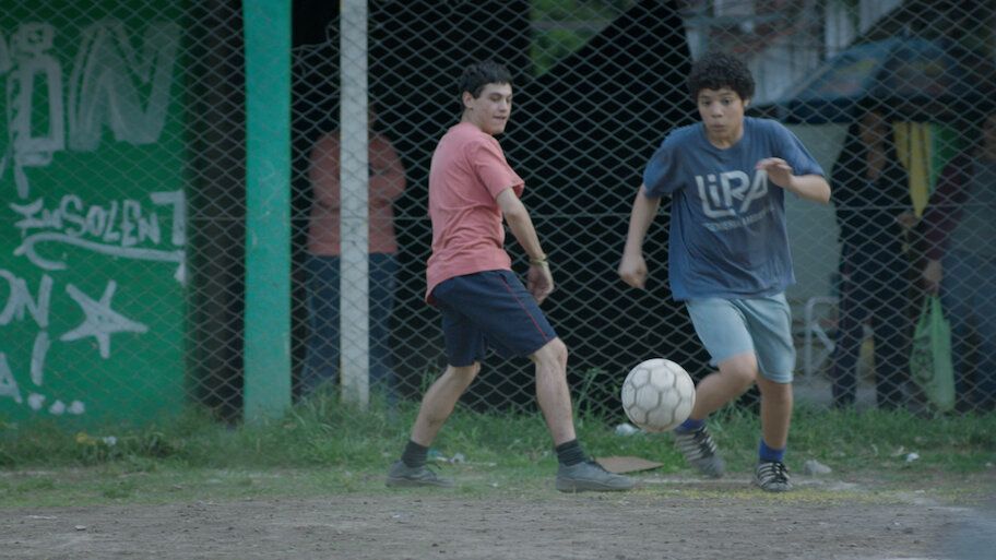 Dos niños juegan al fútbol en un campo verde. Uno con camiseta roja, el otro con azul, ambos concentrados en el balón. 