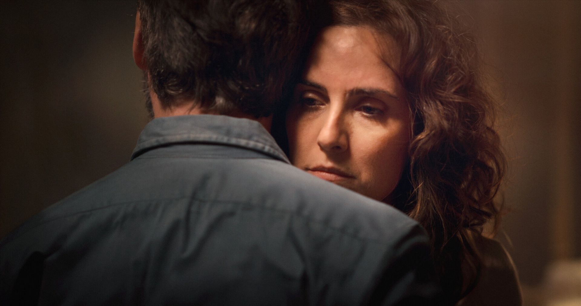 Una mujer de cabello castaño abraza a un hombre, luciendo preocupada en un entorno oscuro.