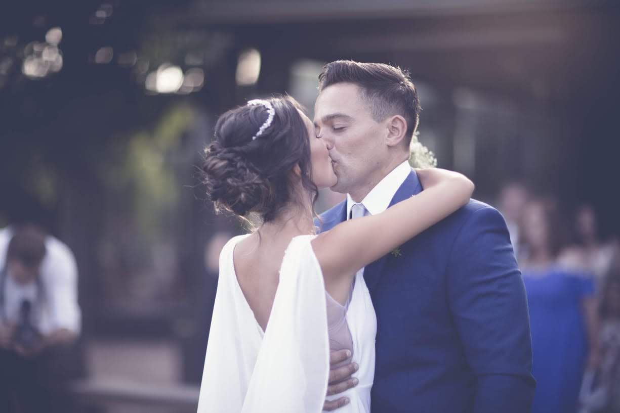 Pareja de novios besándose, abrazándose; novia con vestido blanco, novio con traje azul, al aire libre.
